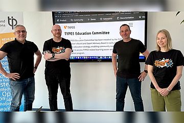 Four people standing in front of a screen displaying the TYPO3 Education Committee webpage. They are inside a room with light-colored walls and a blue banner partially visible on the left. All are casually dressed and smiling at the camera.